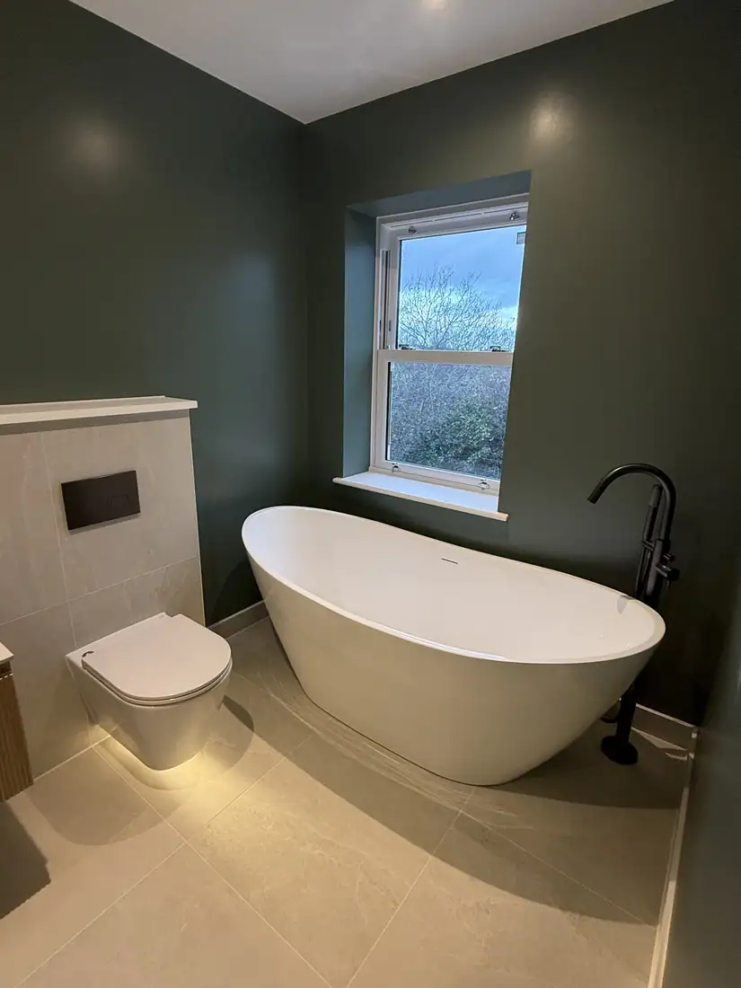 Freestanding bath beneath a window in a green bathroom with a wall-hung WC